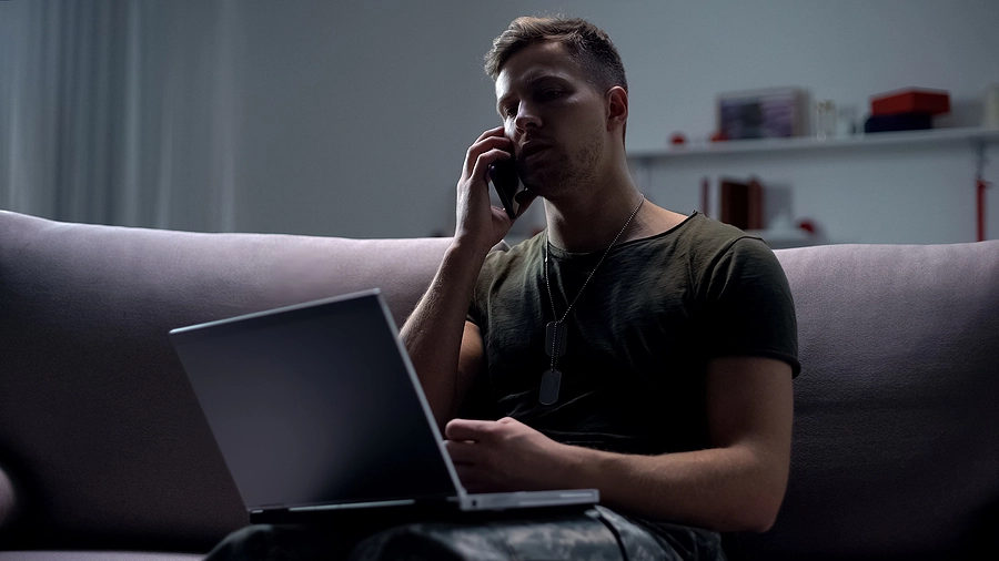 Frustrated veteran sitting on his couch with his laptop talking on the phone to the VA about his disability benefits