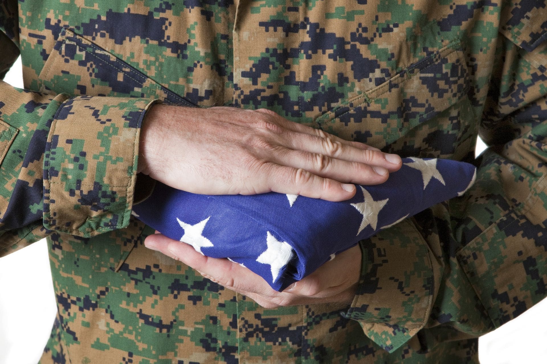 A person in a military camouflage uniform holding a neatly folded American flag with both hands, symbolizing honor, respect, and remembrance.