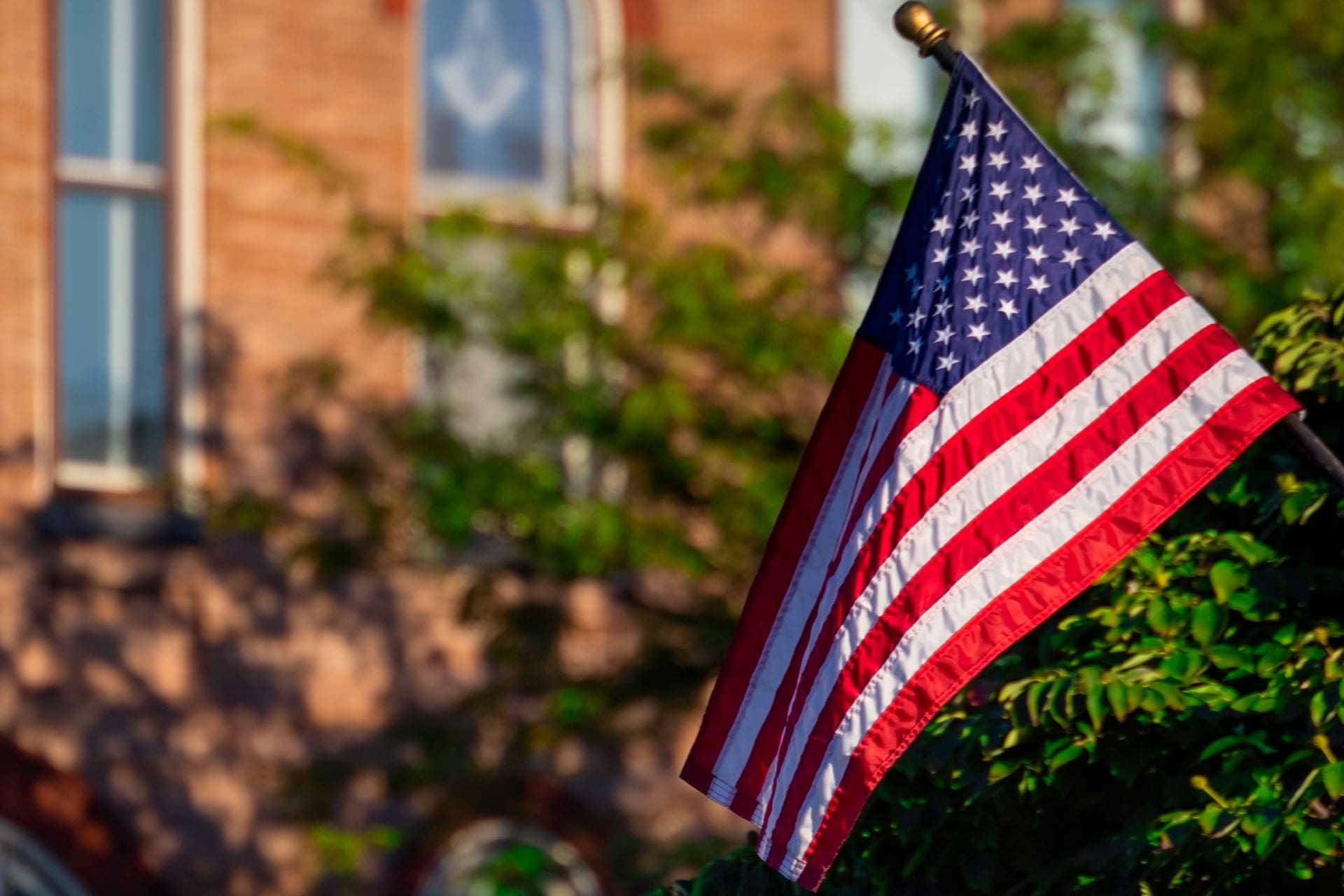 An American flag waving on a flagpole outside a brick building, with green trees in the background, symbolizing patriotism and national pride.